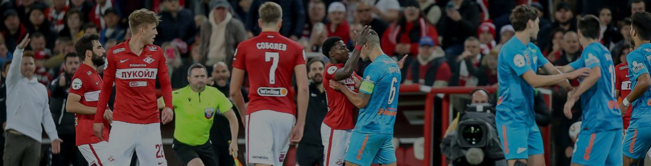 На изображении футболисты в красной и синей форме на поле. Судья в жёлтом. Вокруг спортивная публика. Видно эмоциональные обсуждения среди игроков