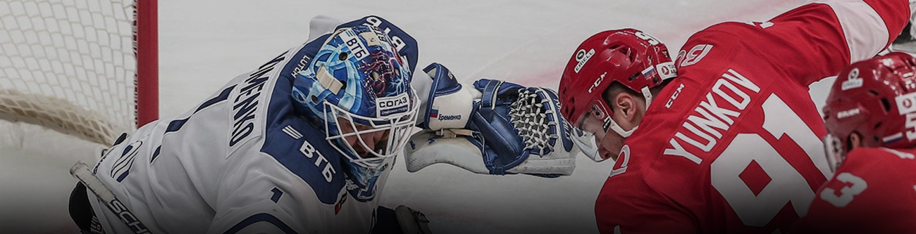 На изображении хоккейный вратарь в бело-синей форме защищает ворота от игрока в красной форме, который атакует. Вратарь ловит шайбу в ловушку близко к льду. Видна активная борьба на льду