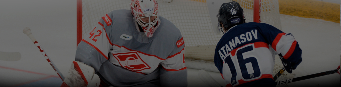 На изображении хоккейный матч: вратарь в светло-серой форме сдерживает шайбу, с игроком команды соперника в тёмно-синей форме, который пытается атаковать. Видны их номера на форме