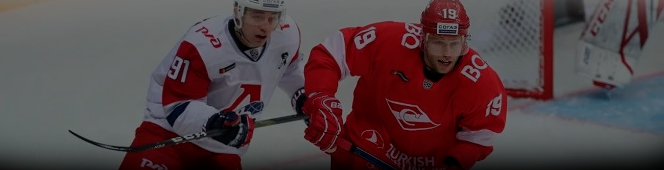 На изображении два хоккеиста на льду в момент игры. Один в красной форме с номером 19, другой в бело-красной с номером 91. Оба сосредоточены на борьбе за шайбу