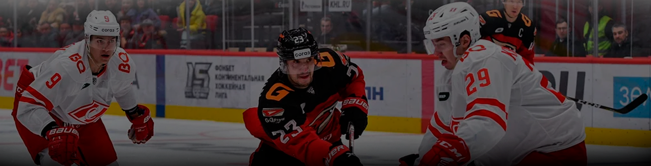 На изображении хоккейный матч: два игрока в бело-красной форме, один в черно-оранжевой. Они борются за шайбу на льду. На заднем плане - зрители за стеклом