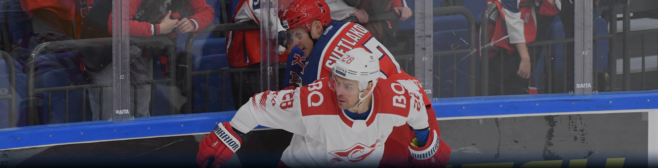 На изображении два хоккеиста борются за шайбу на катке. Один в белой форме с красными элементами, другой - в синей с красным. На заднем плане видны люди за ограждением