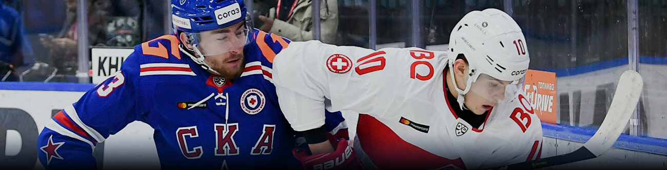 На изображении два хоккеиста участвуют в игре. Игрок в синей форме с эмблемами СКА борется за шайбу с соперником в бело-красной форме. Оба активно двигаются по ледовому покрытию